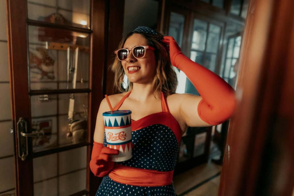 A stylish woman in vintage attire holding a drink indoors, exuding glamour and joy.